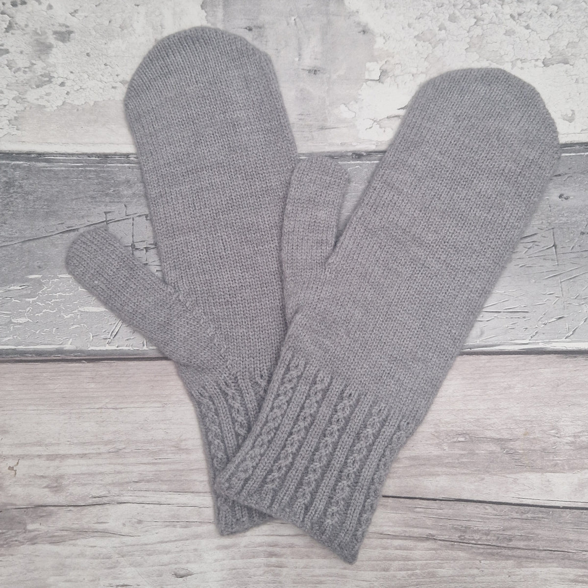A pair of gray knit fingerless mittens with ribbed cable-knit cuffs, displayed on a rustic light gray wooden surface.