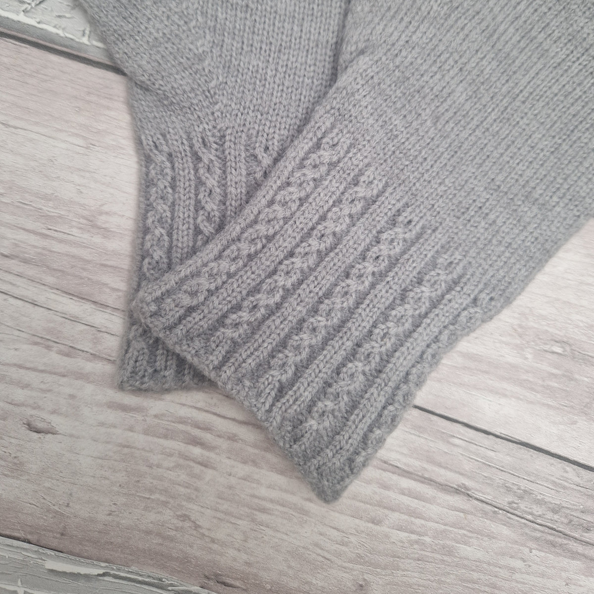 A close-up of a light gray knitted glove, showing the ribbed cuff and cable-knit texture, laid flat on a light gray wood plank surface.