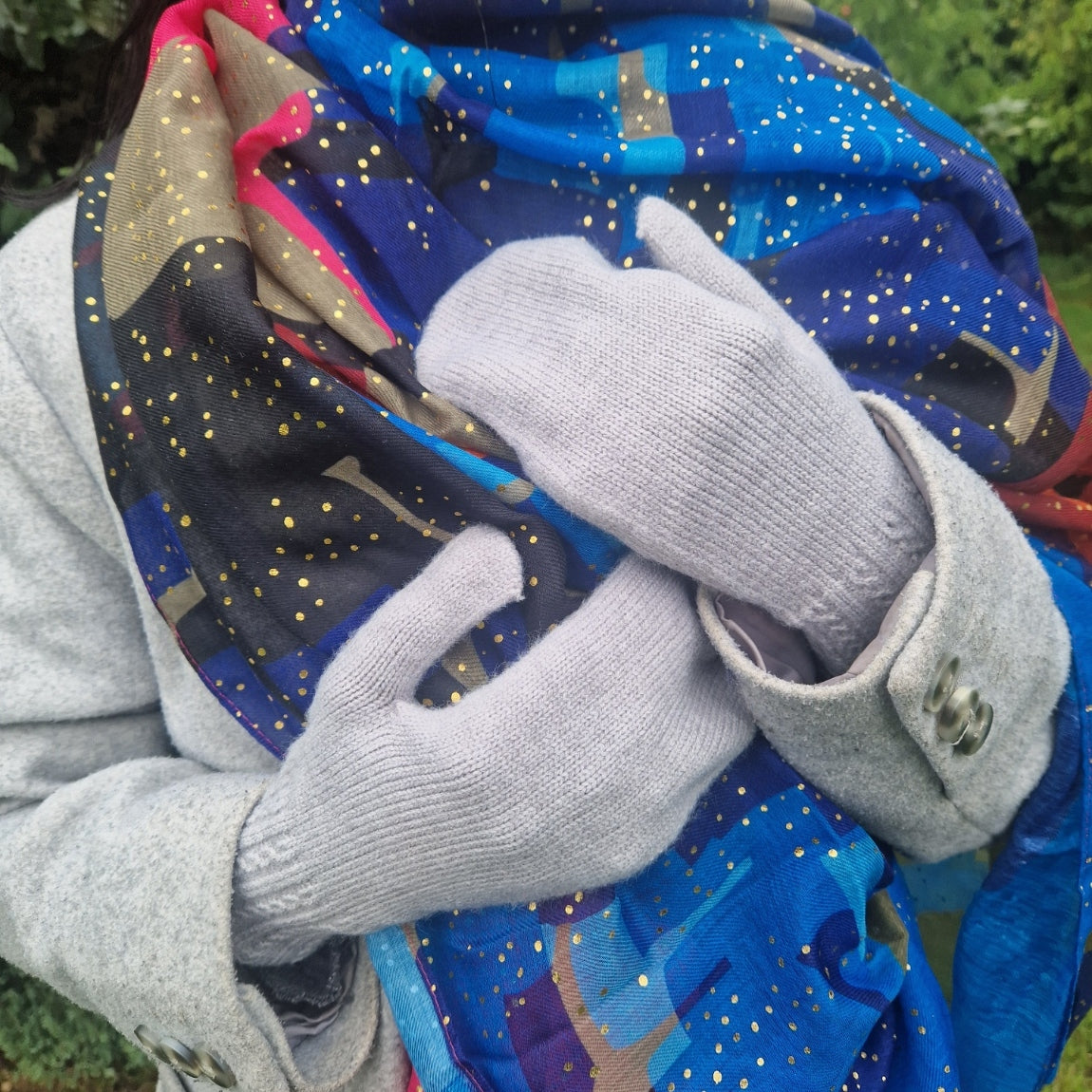 A person outdoors wearing light gray knit gloves and holding a vibrant blue, gold, and black patterned scarf.