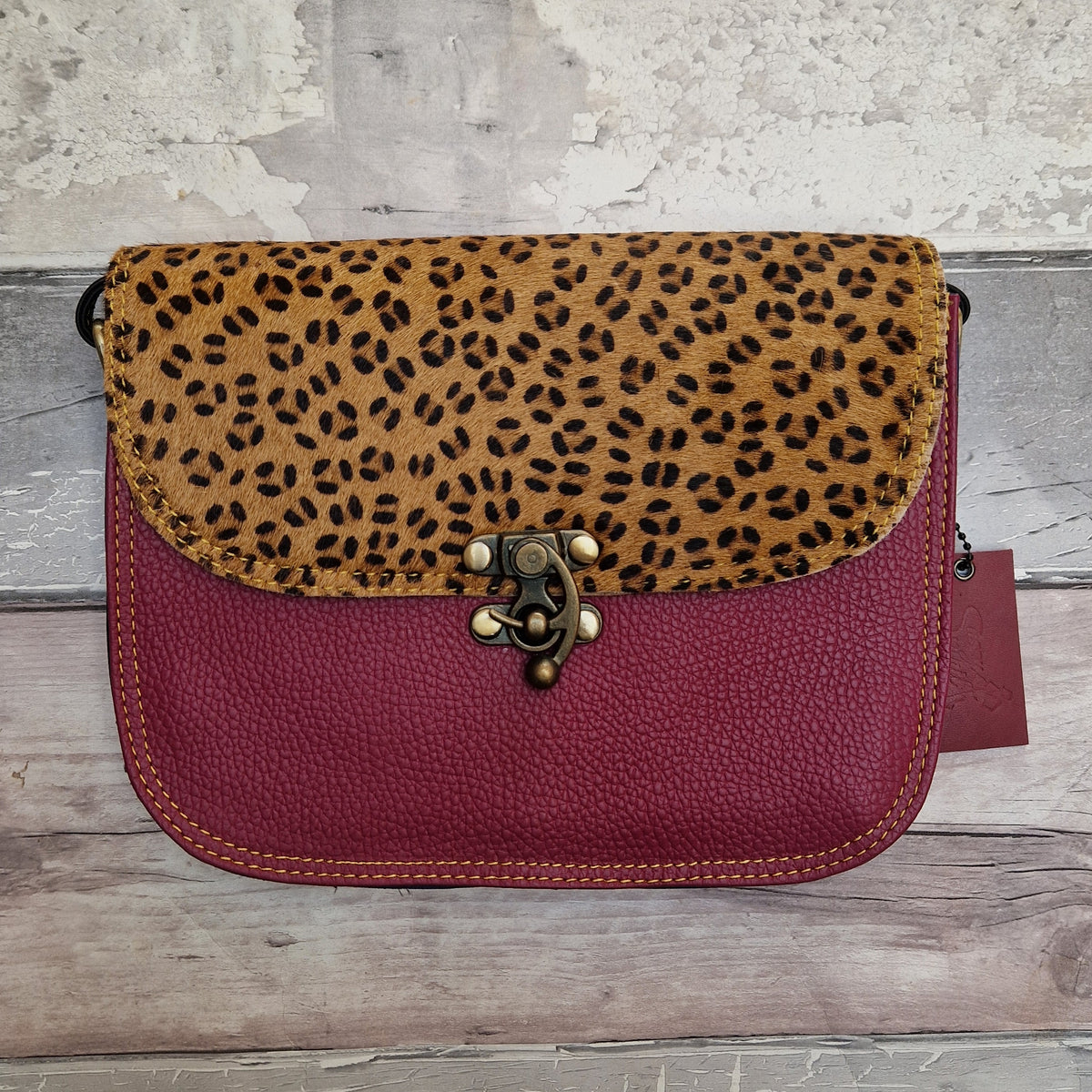 A small shoulder bag with a leopard-print flap and a burgundy pebbled leather body, featuring a decorative metal clasp closure, photographed against a rustic wood and plaster background.