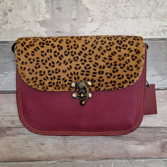 A small shoulder bag with a leopard-print flap and a burgundy pebbled leather body, featuring a decorative metal clasp closure, photographed against a rustic wood and plaster background.