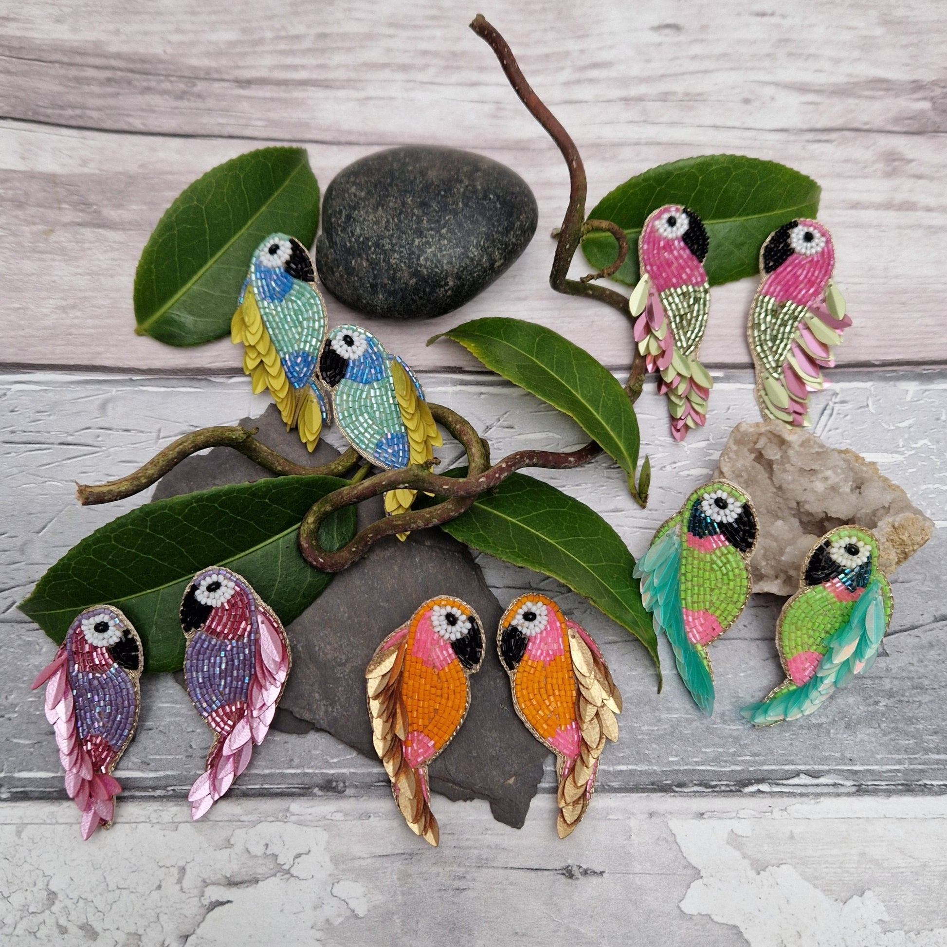 5 Pairs of parrot shaped earrings made from bright coloured beads, each pair in a different colour. Displayed together with leaves and natural materials.