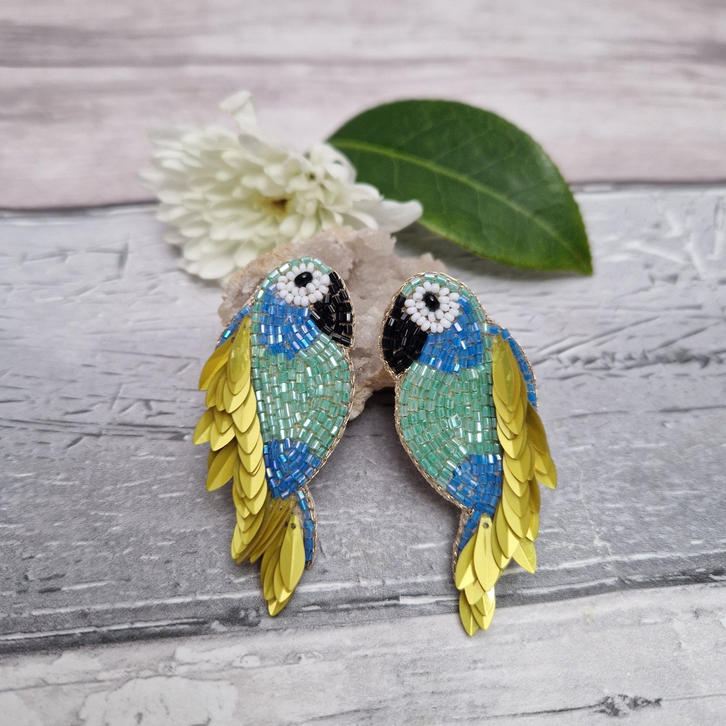 Pair of  blue and green parrot-shaped earrings on a textured surface with a flower and leaf.