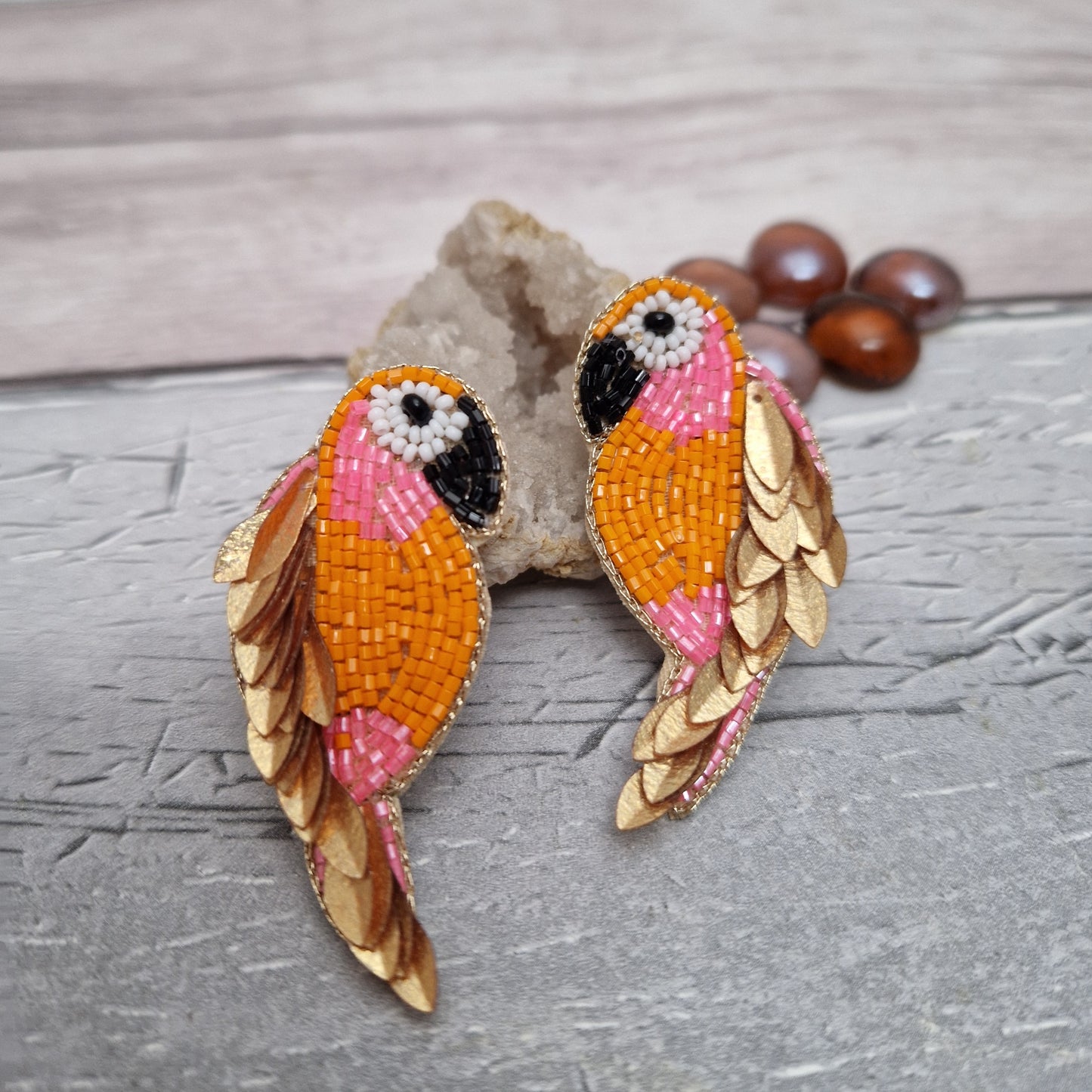 Colorful orange and gold parrot-shaped earrings on a textured surface with beads in the background