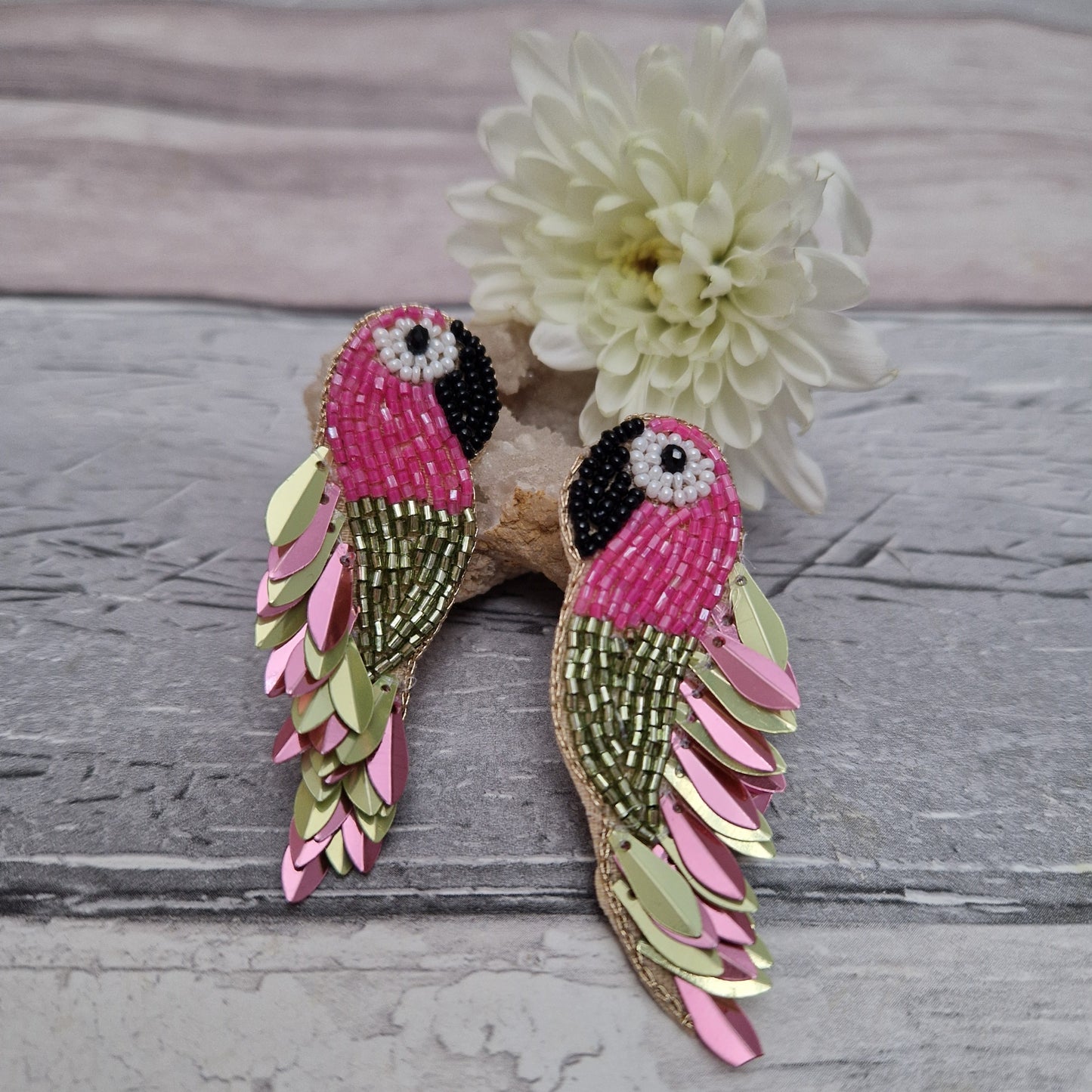 Pair of pink and green parrot-shaped earrings on a wooden surface with a white flower.