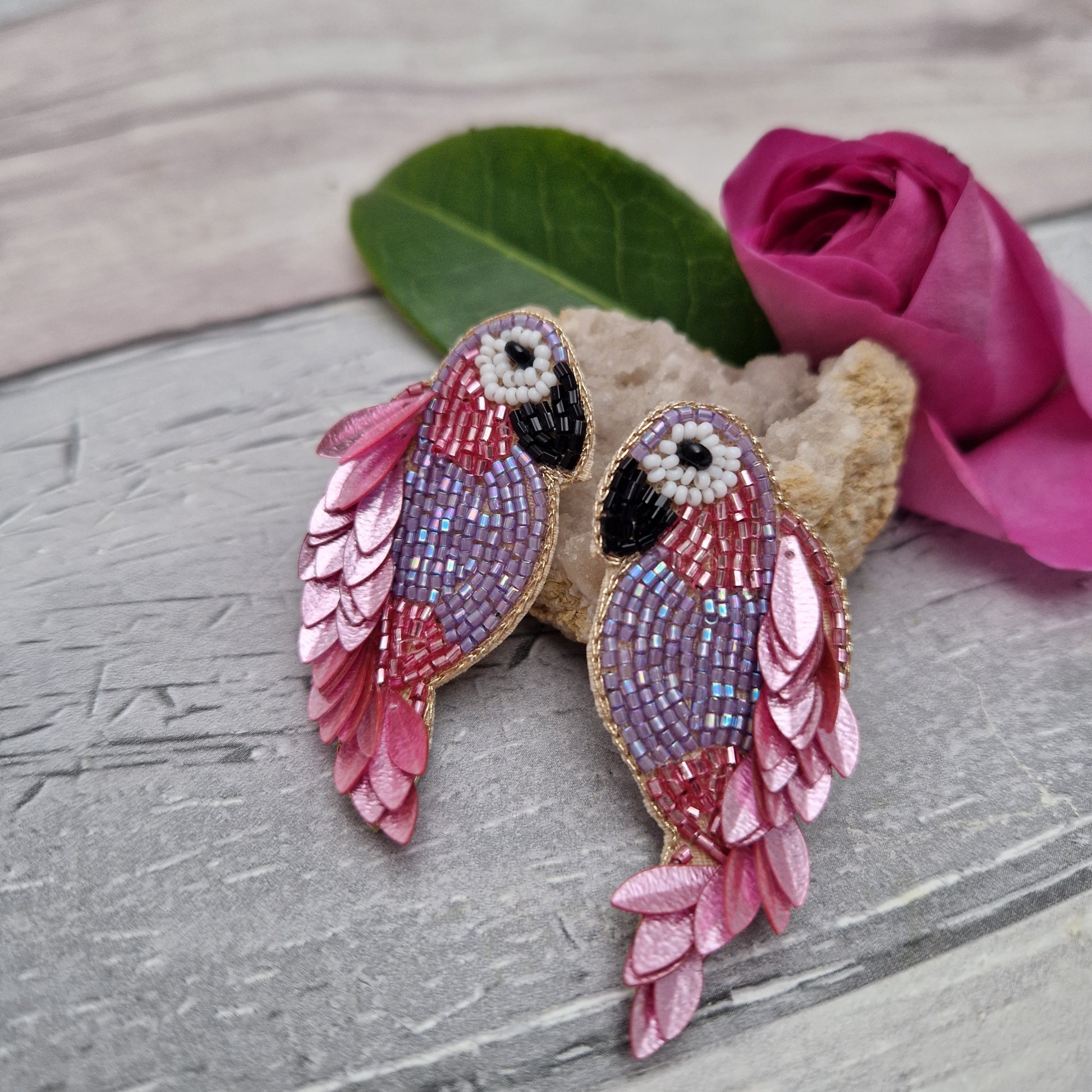 Pair of pink and purple parrot-shaped earrings on a wooden surface with a pink rose.