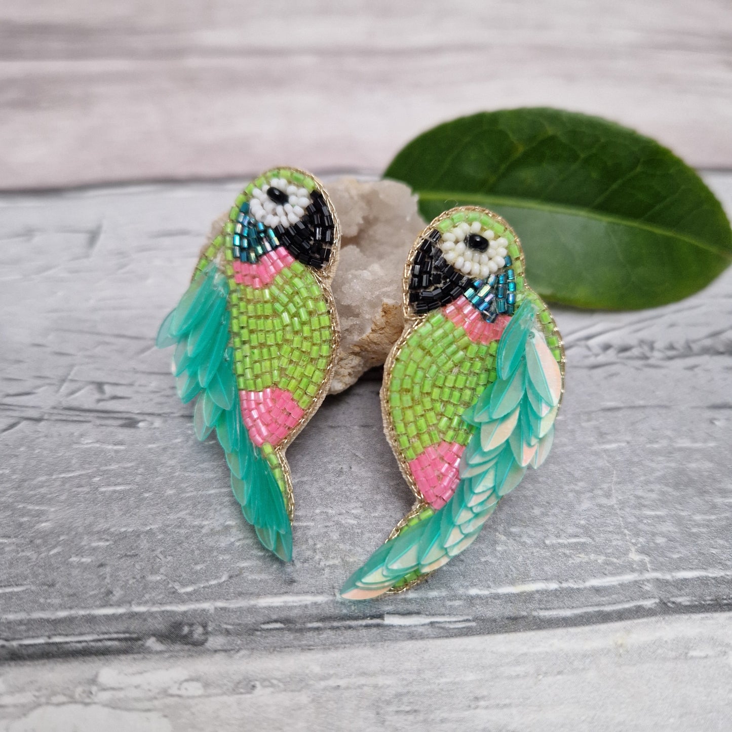 Colorful of mint and pink parrot-shaped earrings on a textured surface with a leaf.