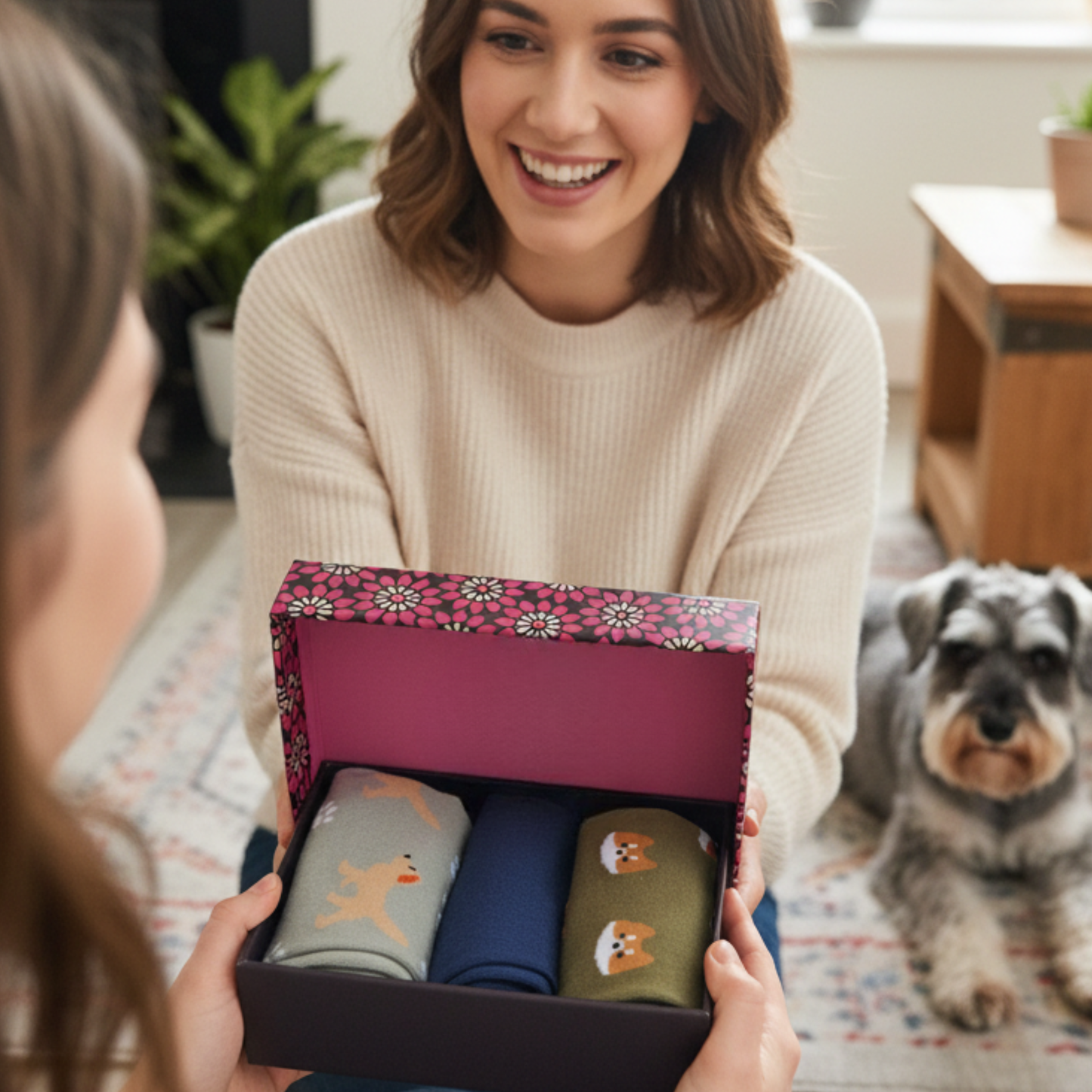 Woman holding a box of socks with a dog design, smiling, with a dog sitting on the floor in the background.