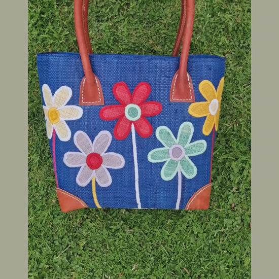 Blue raffia basket bag decorated with long stemmed flowers in a mix of colours. Drawstring top demonstration.