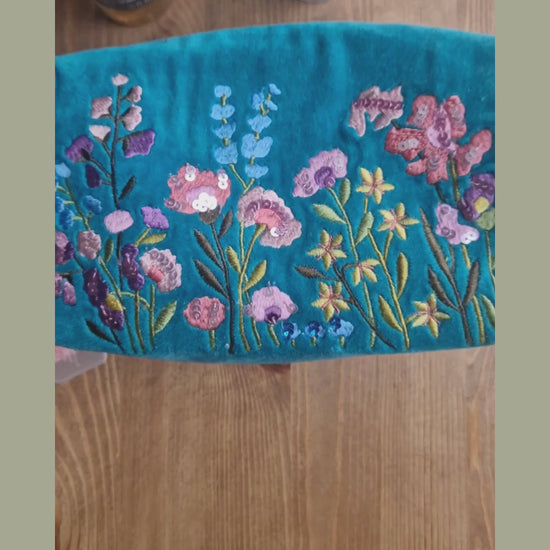Video depicting velvet turquoise wash bag decorated with embroidered flower garden design being loaded with toiletries.